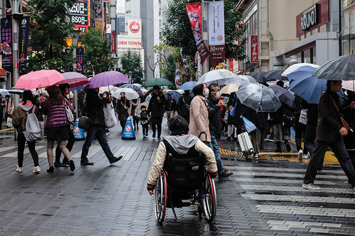 이미지의 필요한 부분만 크롭핑 했음에도 불구하고 고화소로 인해 이미지가 선명하다. 비가 오는 이케부쿠로 길에서 모두 우산을 쓰고 걸어가고 있다. 다리가 불편하신 분이 휠체어 때문에 우산을 쓰지 못하는 모습을 담았다.